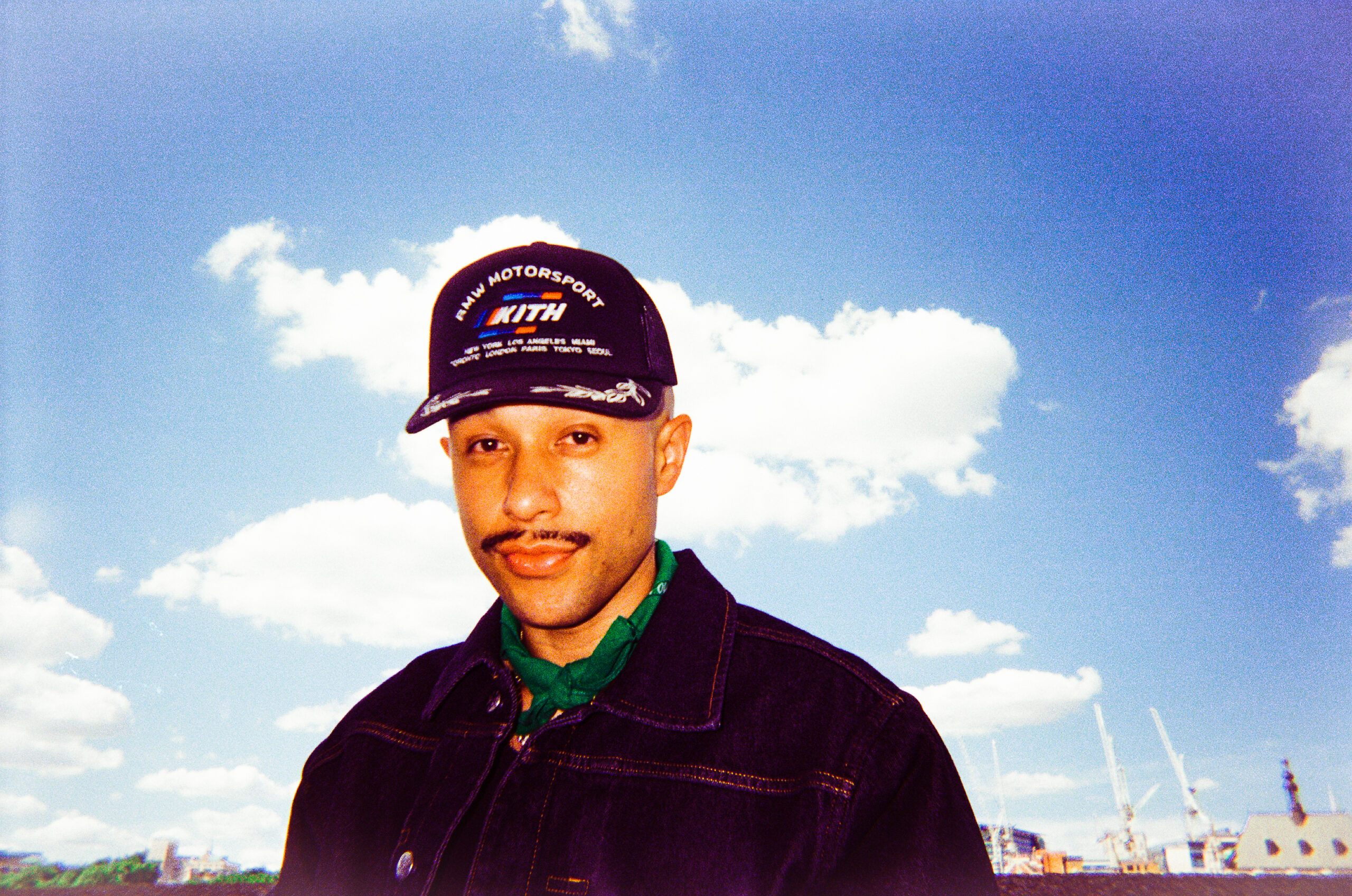 Portrait of a man wearing a dark denim jacket and green scarf, outdoors with a bright blue sky and clouds behind him.
