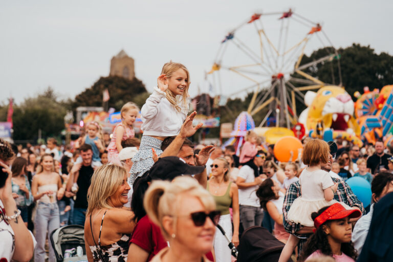 A smiling girl sits on a man's shoulders above a crowded fairground with a Ferris wheel in the background.
