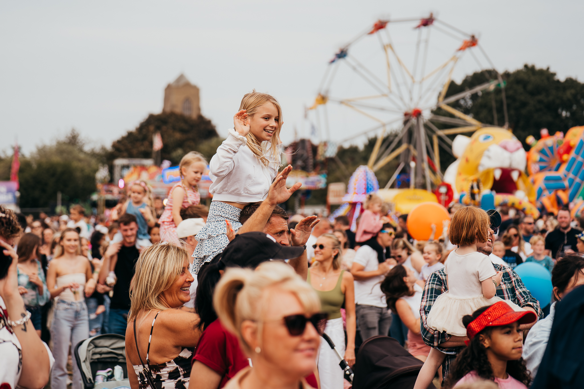 A smiling girl sits on a man's shoulders above a crowded fairground with a Ferris wheel in the background.
