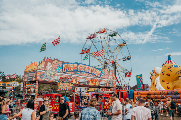 Busy carnival scene with a colorful Ferris wheel, a 'Fire Station' ride facade, and a crowd of people enjoying the fair.