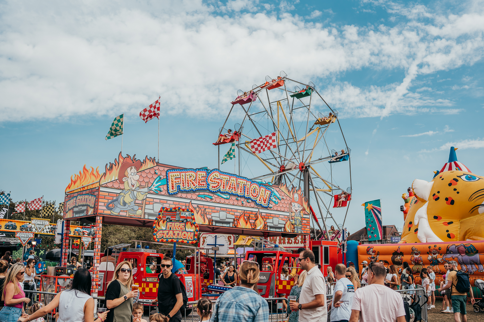 Busy carnival scene with a colorful Ferris wheel, a 'Fire Station' ride facade, and a crowd of people enjoying the fair.